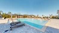 Located at Colonial Gateway Drive in Fort Myers, Florida, this outdoor community pool facility offers a resort-inspired experience designed for relaxation and recreation. The centerpiece is a large, crystal-clear swimming pool with a zero-entry design, surrounded by an expansive stone-paver sun deck that provides ample space for lounging. Rows of wooden and modern chaise lounges are positioned throughout the deck, while pergola structures and private cabanas offer shaded retreats from the Florida sun.

The adjacent clubhouse building features a classic white board-and-batten exterior accented with stone-clad base columns and a pitched roof with dark shingles. A covered outdoor patio extends from the clubhouse, complete with ceiling fans, a ping-pong table, and comfortable seating areas ideal for casual gatherings. An outdoor kitchen and bar seating area is also accessible from the patio.

Restroom facilities and a water fountain station are conveniently located near the pool deck, ensuring practicality for guests. The entire complex is enclosed by a black metal fence, providing both security and a clean aesthetic. Lush natural tree lines frame the background, enhancing the open-air ambiance beneath the abundant Florida sunshine.
