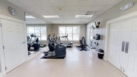 This fitness center offers a clean, functional environment designed to support a well-rounded workout routine. The space is outfitted with a variety of cardio equipment, including treadmills and elliptical machines, arranged to allow comfortable movement throughout the room. A dedicated strength training area features a weight bench and additional exercise equipment, providing options for both cardio and resistance training. Large windows line the walls, flooding the space with natural light and offering views of the surrounding greenery, creating an open and energizing atmosphere. Full-length mirrors along one wall enhance the sense of space while allowing users to monitor their form during exercise. A wall-mounted flat-screen TV adds an entertainment element to workouts. The room features a light-toned, easy-to-clean hard floor surface and a drop ceiling with recessed lighting panels that ensure consistent, even illumination throughout. Neutral wall tones and a tidy layout contribute to a calm and focused environment. The fitness center is a single-level facility that is straightforward to navigate, making it accessible and welcoming for users of all fitness levels.