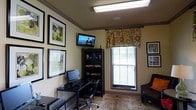 This single-floor home office is thoughtfully arranged to balance productivity and comfort. The room features warm, neutral-toned walls accented by gray crown molding and a tray ceiling detail, lending a polished architectural finish to the space. Overhead, a flush fluorescent light fixture provides ample illumination throughout the room.

The workspace is anchored by a compact desk paired with a rolling office chair and a desktop computer setup, positioned conveniently against one wall. Adjacent to the desk, a dark bookcase offers organized storage for books, supplies, and personal items. A wall-mounted flat-screen television is installed above the bookcase, adding a multimedia element to the room.

A single window dressed with a vibrant floral valance in green, orange, and pink tones introduces a pop of color and personality, while slatted blinds allow for light control and privacy. The window draws in natural light, brightening the space effectively. Decorative framed artwork adorns the surrounding walls, enhancing the room's character. A plush armchair positioned in the corner provides an additional seating option, and a patterned area rug grounds the overall layout with visual warmth.