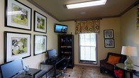 This single-floor home office is thoughtfully arranged to balance productivity and comfort. The room features warm, neutral-toned walls accented by gray crown molding and a tray ceiling detail, lending a polished architectural finish to the space. Overhead, a flush fluorescent light fixture provides ample illumination throughout the room.

The workspace is anchored by a compact desk paired with a rolling office chair and a desktop computer setup, positioned conveniently against one wall. Adjacent to the desk, a dark bookcase offers organized storage for books, supplies, and personal items. A wall-mounted flat-screen television is installed above the bookcase, adding a multimedia element to the room.

A single window dressed with a vibrant floral valance in green, orange, and pink tones introduces a pop of color and personality, while slatted blinds allow for light control and privacy. The window draws in natural light, brightening the space effectively. Decorative framed artwork adorns the surrounding walls, enhancing the room's character. A plush armchair positioned in the corner provides an additional seating option, and a patterned area rug grounds the overall layout with visual warmth.