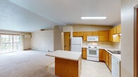 This single-floor residence offers a well-organized and functional layout with a warm, neutral color palette throughout. The open-concept kitchen is a central feature, outfitted with honey-toned wood cabinetry, white appliances including a refrigerator, electric range, microwave, and dishwasher, and a center island with a light countertop that provides additional workspace and storage. Light tile flooring runs through the kitchen, transitioning to plush carpet in the living and bedroom areas. The living area benefits from a vaulted ceiling that adds a sense of openness and volume to the space, along with a ceiling fan for year-round comfort. Baseboard heating units are present throughout, ensuring consistent warmth across the home. The bedrooms are generously sized, each featuring ceiling fans, large windows that invite natural light and outdoor views, and plush carpeting. One bedroom includes a spacious walk-in closet with wire shelving and hanging rods for ample storage. The full bathroom is finished with warm wood cabinetry, a wide vanity with a single sink, a large mirror with overhead lighting, and light tile flooring. Additional closet space and a practical hallway layout contribute to the overall functionality of this well-appointed home.