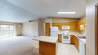 This single-floor residence offers a well-organized and functional layout with a warm, neutral color palette throughout. The open-concept kitchen is a central feature, outfitted with honey-toned wood cabinetry, white appliances including a refrigerator, electric range, microwave, and dishwasher, and a center island with a light countertop that provides additional workspace and storage. Light tile flooring runs through the kitchen, transitioning to plush carpet in the living and bedroom areas. The living area benefits from a vaulted ceiling that adds a sense of openness and volume to the space, along with a ceiling fan for year-round comfort. Baseboard heating units are present throughout, ensuring consistent warmth across the home. The bedrooms are generously sized, each featuring ceiling fans, large windows that invite natural light and outdoor views, and plush carpeting. One bedroom includes a spacious walk-in closet with wire shelving and hanging rods for ample storage. The full bathroom is finished with warm wood cabinetry, a wide vanity with a single sink, a large mirror with overhead lighting, and light tile flooring. Additional closet space and a practical hallway layout contribute to the overall functionality of this well-appointed home.