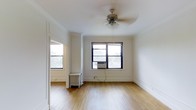 This single-floor residence blends classic architectural charm with functional living. The main living area is finished with warm light hardwood flooring that extends throughout, creating a cohesive and inviting atmosphere. Crisp white walls and crown molding add a timeless, prewar character to the space, while large black-framed windows on multiple sides flood the room with generous natural light and offer views of the surrounding neighborhood.

A ceiling fan with a light fixture is centrally mounted, providing both ventilation and ambient lighting. The room is equipped with a wall-mounted air conditioning unit, complemented by a traditional cast-iron radiator, ensuring year-round comfort through both warm and cold seasons. The clean, open layout of the living area allows for versatile furniture arrangements and a sense of spaciousness.

Doorways connect seamlessly to additional areas of the unit, hinting at a well-organized floor plan. The combination of original architectural details — from the decorative moldings to the classic radiator — and practical modern amenities gives this space a distinctive character that balances historic appeal with everyday functionality.