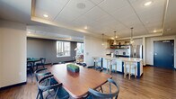 Located at 14211 Akron Ave in Rosemount, MN, this single-floor communal space offers a thoughtfully designed open-plan layout that seamlessly connects a living room, dining room, and kitchen. The interior features warm wood-look flooring throughout, a drop tile ceiling with recessed lighting, and a neutral color palette accented by deep charcoal feature walls.

The kitchen is fully equipped with stainless steel appliances, including a gas range with a stainless steel hood vent, a dishwasher, and a full-size refrigerator. Dark cabinetry is paired with white countertops and a subway tile backsplash, while open shelving adds a modern touch. A central island with bar seating and pendant lighting enhances both function and style.

The dining area accommodates a large wooden table with multiple chairs, making it ideal for group gatherings. Large windows flood the space with natural light and offer views of the surrounding landscape. Framed artwork and greenery add warmth to the area.

The living room features a comfortable sofa, accent chair, and oval coffee table, arranged around a wall-mounted television. A large window overlooks an outdoor patio, bringing in abundant daylight. Textured wallcovering and layered area rugs complete the inviting, residential ambiance of this versatile shared space.