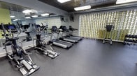 This fitness center is thoughtfully designed to provide a functional and motivating workout environment. The space is outfitted with a lineup of modern cardio equipment, including multiple treadmills with integrated display screens, an elliptical trainer, and a rowing machine, offering a diverse range of aerobic exercise options. Rubber flooring throughout the gym provides durability and safety underfoot, ideal for both cardio and strength training activities. A full-length mirror spans one wall, enhancing the sense of space while allowing users to monitor their form during workouts. Industrial-style ceiling-mounted lighting ensures the area is well-lit, complemented by wall-mounted fans that support air circulation during intense sessions. A striking decorative lattice partition with a gold-toned geometric pattern serves as a distinctive design element, adding visual interest to the otherwise utilitarian setting. Free weights and additional strength training equipment are also available, making this a versatile facility. The overall ambiance is clean, practical, and purposeful, catering to a variety of fitness routines in a compact yet efficiently organized layout.
