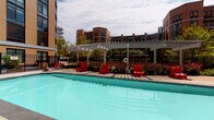 This single-floor space includes a half bathroom and access to a well-appointed outdoor pool area set within a multi-story brick residential complex. The half bathroom is finished with warm beige walls, a wood-framed vanity topped with a white countertop and integrated sink, chrome fixtures, a brushed wood-framed mirror, and an oil-rubbed bronze towel ring—all contributing to a classically understated aesthetic. A white door with a brass knob provides privacy, and wood flooring is visible at the entry threshold.

The outdoor pool area serves as a central communal amenity. The generously sized rectangular pool features clear turquoise water and is framed by a spacious tiled deck lined with bold red lounge chairs and seating arrangements. A white pergola structure anchors the space, providing shaded gathering areas beneath an open-lattice canopy. Lush landscaping, including flowering shrubs and mature trees, borders the perimeter, while a wrought iron fence encloses the area. The surrounding multi-story brick buildings frame the courtyard, creating a defined and cohesive urban outdoor environment bathed in natural light.