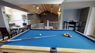 This single-floor entertainment space makes a strong impression with its striking vaulted ceiling clad in warm wood paneling, angled beams, and recessed track lighting that casts a focused glow across the room. The centerpiece is a full-size pool table with a vibrant blue felt surface, perfectly positioned beneath the soaring ceiling to allow ample room for play. A ceiling fan adds both function and a casual ambiance to the space.

The open layout is defined by a split-level design, with a railed landing area that overlooks a lower section, adding architectural depth and visual interest. The walls are finished in a contemporary dark gray tone, creating a moody, lounge-like atmosphere that complements the warm wood overhead. A corner seating area with a television provides a relaxed retreat for spectators, while a tall decorative plant adds a natural element to the room.

Adjacent to the pool table, a dark upright piano offers a refined counterpoint to the recreational energy of the space. The combination of entertainment features, architectural character, and thoughtful design details makes this a versatile and inviting area well-suited for socializing and leisure.
