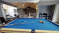 This single-floor entertainment space makes a strong impression with its striking vaulted ceiling clad in warm wood paneling, angled beams, and recessed track lighting that casts a focused glow across the room. The centerpiece is a full-size pool table with a vibrant blue felt surface, perfectly positioned beneath the soaring ceiling to allow ample room for play. A ceiling fan adds both function and a casual ambiance to the space.

The open layout is defined by a split-level design, with a railed landing area that overlooks a lower section, adding architectural depth and visual interest. The walls are finished in a contemporary dark gray tone, creating a moody, lounge-like atmosphere that complements the warm wood overhead. A corner seating area with a television provides a relaxed retreat for spectators, while a tall decorative plant adds a natural element to the room.

Adjacent to the pool table, a dark upright piano offers a refined counterpoint to the recreational energy of the space. The combination of entertainment features, architectural character, and thoughtful design details makes this a versatile and inviting area well-suited for socializing and leisure.