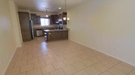 This two-story home offers a well-appointed layout designed for comfortable everyday living. The main level is anchored by an open-concept kitchen and living area that seamlessly connects the two spaces. The kitchen is a standout feature, showcasing rich dark wood cabinetry with glass-front upper panels, granite countertops, and stainless steel appliances including a dishwasher and refrigerator. A kitchen peninsula provides additional workspace and casual seating potential, while a decorative wrought iron chandelier and recessed pendant lighting add warmth and character overhead. Natural light filters in through a window above the sink, brightening the space. The generous open area adjacent to the kitchen is finished with large-format beige ceramic tile flooring that extends throughout the main level, creating a cohesive and easy-to-maintain surface. Neutral wall tones complement the warm wood tones of the cabinetry, giving the space a welcoming and versatile ambiance. The two-floor layout offers additional living or sleeping accommodations on the upper level, providing a practical separation between common and private areas of the home.