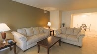 This single-floor residence offers a comfortable and thoughtfully arranged living environment. The living room serves as the centerpiece of the space, featuring a neutral color palette that creates a calm and welcoming atmosphere. A textured ceiling adds subtle visual interest overhead, while the walls are finished in a combination of warm olive green and soft white tones, providing a stylish contrast that anchors the room. Plush, light-toned sofas are arranged around a dark wood coffee table, creating a functional and inviting seating area ideal for relaxing or entertaining. A matching end table with a classic table lamp adds a warm glow to the space, enhancing its cozy feel. The flooring is covered in wall-to-wall carpet, contributing to the overall sense of comfort underfoot. A decorative plant near the hallway entrance adds a touch of greenery, while an open doorway visible in the background suggests a smooth and accessible flow between rooms. The layout is practical and open, making efficient use of the available space on a single level.