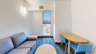 Located at 304 College Ave in Ithaca, NY, this two-bedroom, one-bathroom unit offers a thoughtfully designed, modern interior across a single floor. The open-concept kitchen and living area serves as the heart of the unit. The kitchen features stainless steel appliances—including an electric range, dishwasher, and over-the-range microwave—paired with light wood cabinetry, granite-style countertops, and wood-look flooring that gives the space a clean, contemporary feel.

The living area transitions seamlessly from the kitchen, featuring blue-toned carpet tiles, wall-mounted lighting, and a window with outdoor views of the surrounding Ithaca neighborhood. A dining nook with a round table and chairs provides a functional space for meals or study.

Both bedrooms are carpeted and feature natural light from windows with roller shades. Each room is furnished with a bed, a light wood dresser, and a dedicated study desk, making them well-suited for student living. The primary bedroom is comfortably sized, offering room for essential furnishings.

The shared bathroom is clean and functional, featuring light wood cabinetry, a granite-top vanity, a walk-in shower with glass enclosure and grab bars, and wood-look tile flooring. The overall design is modern, neutral, and practical throughout.