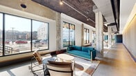 Located on History Walk in Baltimore's vibrant Fells Point neighborhood (21231), this single-floor commercial space offers a generously sized, open-plan layout that blends raw industrial character with thoughtfully curated modern design. The space is defined by its exposed concrete structural columns, which rise to meet a distinctive textured ceiling fitted with track lighting and visible ductwork — hallmarks of adaptive reuse architecture. Large, floor-to-ceiling windows line the street-facing wall, flooding the interior with abundant natural light and offering open views of the surrounding urban streetscape. Colorful geometric window graphics add a bold, contemporary visual element to the façade. The polished and raw concrete flooring throughout reinforces the industrial aesthetic while providing a neutral canvas that accommodates a variety of uses. A lounge area with modern seating and a separate dining or meeting table configuration demonstrate the space's versatility. Toward the rear, light blue accent walls, decorative murals, and large padded utility doors introduce color and artistic character. Lush potted plants are thoughtfully placed throughout, softening the industrial elements and adding warmth. This flexible, light-filled space is well-suited for retail, creative office, or community-facing commercial use.