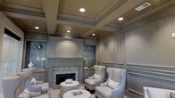 This distinguished single-floor space showcases an exceptional level of architectural craftsmanship and refined interior design. The centerpiece of the main living area is a classic fireplace framed by intricate mosaic tile detailing and flanked by custom built-in cabinetry with display niches, ideal for showcasing decorative accents and collectibles. The walls are dressed in elegant raised panel millwork finished in a warm taupe-grey palette, lending a cohesive and polished aesthetic throughout. Above, the coffered ceiling commands attention with its deep, structured grid work and recessed lighting that casts a warm, inviting glow across the room. The seating arrangement features a pair of tall wingback chairs in a soft grey upholstery, complemented by coordinating accent pillows and a rounded pedestal side table, creating an intimate and comfortable conversation area. The overall ambiance is one of understated luxury — thoughtfully designed with attention to detail at every turn. Classic proportions, neutral tones, and layered lighting combine to produce a space that feels both stately and welcoming, making it well-suited for formal entertaining or refined relaxation.