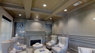 This distinguished single-floor space showcases an exceptional level of architectural craftsmanship and refined interior design. The centerpiece of the main living area is a classic fireplace framed by intricate mosaic tile detailing and flanked by custom built-in cabinetry with display niches, ideal for showcasing decorative accents and collectibles. The walls are dressed in elegant raised panel millwork finished in a warm taupe-grey palette, lending a cohesive and polished aesthetic throughout. Above, the coffered ceiling commands attention with its deep, structured grid work and recessed lighting that casts a warm, inviting glow across the room. The seating arrangement features a pair of tall wingback chairs in a soft grey upholstery, complemented by coordinating accent pillows and a rounded pedestal side table, creating an intimate and comfortable conversation area. The overall ambiance is one of understated luxury — thoughtfully designed with attention to detail at every turn. Classic proportions, neutral tones, and layered lighting combine to produce a space that feels both stately and welcoming, making it well-suited for formal entertaining or refined relaxation.
