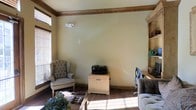 This single-floor space combines comfort and functionality in a welcoming, well-appointed interior. The room is framed by rich, dark wood crown molding that contrasts beautifully against the soft cream-toned walls, lending a classic and refined character to the space. Hardwood floors run throughout, adding warmth and a sense of continuity underfoot.

Natural light filters generously through multiple windows fitted with wooden blinds, casting a bright and inviting atmosphere across the room. The layout thoughtfully accommodates both a relaxed seating area and a dedicated workspace. A tufted wingback chair anchors one corner, providing a classic and comfortable spot for reading or conversation, while a built-in shelving unit along one wall offers organized storage and display space.

The home office area features a desk and task chair positioned near the built-in shelves, making it an efficient and integrated workspace. A compact storage cabinet serves as a practical surface, currently hosting a multifunction printer. Decorative touches, including potted greenery and natural accents, add life and texture to the room.

Overall, the space strikes a balance between traditional design elements and practical living, making it a versatile and inviting environment suited for both work and leisure.