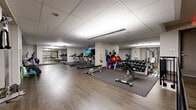 This fitness center offers a comprehensive workout environment spread across a single, open floor plan. The space features durable wood-look flooring throughout, a drop tile ceiling with recessed and linear lighting, and full-length mirrored walls that visually expand the room and allow users to monitor their form during exercise.

The cardio zone is anchored by a row of treadmills positioned in front of a wall-mounted flat-screen television, making for an engaging workout experience. Complementing the treadmills are an elliptical trainer and a recumbent stationary bike, providing a well-rounded selection of low- and high-impact cardio options.

The strength training area is equally well-appointed, featuring a tiered dumbbell rack stocked with a wide range of free weights, adjustable benches, and a multi-station cable machine suitable for a variety of resistance exercises. Functional fitness equipment including stability balls, resistance bands, and kettlebells rounds out the offerings.

An open stretching and floor exercise area provides ample space for warm-ups, cool-downs, and bodyweight training. Rubber flooring mats are placed strategically beneath equipment zones for added safety and protection. The space is accessible via a railed staircase entrance, and emergency exit signage is clearly posted throughout, ensuring a safe and organized environment for all users.