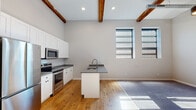 Located on Harris Avenue in Providence's West End, this single-floor loft-style unit showcases a compelling blend of industrial heritage and contemporary design. Soaring ceilings with exposed wooden beams and visible piping set a distinctive architectural tone throughout the open living and kitchen area, while warm hardwood flooring anchors the space with natural texture.

The kitchen is well-appointed with white shaker cabinetry extending to ceiling height, stainless steel appliances including a refrigerator, range, and microwave, and a central island with a gray countertop and integrated sink. A wall of large, light-filtering windows floods the space with natural light, complementing the open layout.

Bedrooms feature plush carpeting, wall-mounted mini-split HVAC units, and ample natural light from generously sized windows with blinds. One bedroom retains a striking exposed brick accent wall, adding warmth and character. Walk-in closet access is also present.

The bathroom offers a dual-sink vanity with gray countertops, white cabinetry with matte black hardware, round mirrors, and elegant tile flooring. Throughout, the space balances its industrial roots with clean, modern finishes for a refined yet characterful living environment.