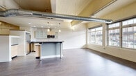 Located at 230 Scott Street in Buffalo, NY, this single-floor loft-style unit showcases an industrial-modern aesthetic with exposed spiral ductwork running along the ceiling and light wood-planked ceiling details that lend a warm, converted-warehouse character. The open-concept living and kitchen area is spacious and airy, with durable hardwood-style flooring throughout and a neutral color palette that enhances the sense of openness.

The kitchen is a standout feature, equipped with stainless steel appliances including a gas range, microwave, and refrigerator. Light maple cabinetry with frosted glass upper panels pairs with dark granite countertops, and a central island with bar seating provides additional prep and dining space. Pendant lighting above the island adds a polished finish.

Large, multi-pane windows along the exterior wall flood the living area with natural light and offer street-level views of the surrounding Buffalo neighborhood. The bedroom area features the same wide-plank flooring and a bold gray accent wall, also benefiting from generous window access.

The bathroom is clean and modern, with a granite-topped vanity, wood-toned cabinetry, and tile flooring. A dedicated utility room houses the furnace and in-unit washer/dryer, providing added convenience. This well-appointed urban unit blends industrial character with contemporary finishes.