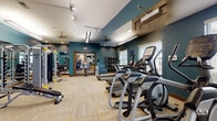 Located at 1998 Persimmon Way in Tipp City, Ohio, this single-floor fitness center is a well-appointed facility designed to support a comprehensive workout experience. The space is defined by bold teal accent walls paired with neutral gray ceilings, patterned carpet tile flooring in the cardio and strength zones, and smooth hardwood-style flooring in the dedicated exercise and stretching area.

The main workout floor is generously sized and features a strong lineup of cardio equipment, including treadmills, elliptical cross-trainers, upright and recumbent stationary bikes, and a cycling/spin bike. A multi-station strength machine, adjustable weight bench, barbell rack, and free weights round out the strength training offerings. Ceiling fans provide additional air circulation throughout the space.

A mirrored wall section creates an open, studio-like area ideal for stretching, balance work, or functional fitness, and is equipped with stability balls, plyo boxes, and resistance training accessories. A wall-mounted flat-screen television adds entertainment options for cardio sessions.

Large floor-to-ceiling windows and a full glass garage-style door flood the space with natural light while offering views of the surrounding outdoor landscape. The entryway features double glass doors, a small cubby storage unit, and a water fountain, ensuring convenient access and practical amenities for users.