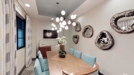 This single-story space showcases a thoughtfully designed dining area that blends contemporary and coastal influences. The room centers around a natural wood dining table with a herringbone-patterned surface, surrounded by upholstered chairs in a distinctive teal geometric print with nailhead trim detailing. A striking multi-globe sputnik chandelier serves as the focal point overhead, suspended from a recessed ceiling panel painted in a soft slate blue — a subtle architectural detail that adds depth and visual interest to the otherwise neutral palette. The walls are finished in a light textured treatment, providing a refined backdrop for an eclectic gallery of organic, geode-inspired wall sculptures in muted silver and stone tones. A wall-mounted flat-screen television and a warm wood sideboard add functional elements to the space. A fresh orchid arrangement at the center of the table introduces a natural touch. The overall ambiance is polished yet inviting, combining clean lines with artful décor to create a dining environment that feels both sophisticated and approachable.