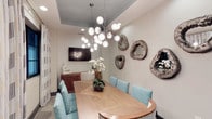 This single-story space showcases a thoughtfully designed dining area that blends contemporary and coastal influences. The room centers around a natural wood dining table with a herringbone-patterned surface, surrounded by upholstered chairs in a distinctive teal geometric print with nailhead trim detailing. A striking multi-globe sputnik chandelier serves as the focal point overhead, suspended from a recessed ceiling panel painted in a soft slate blue — a subtle architectural detail that adds depth and visual interest to the otherwise neutral palette. The walls are finished in a light textured treatment, providing a refined backdrop for an eclectic gallery of organic, geode-inspired wall sculptures in muted silver and stone tones. A wall-mounted flat-screen television and a warm wood sideboard add functional elements to the space. A fresh orchid arrangement at the center of the table introduces a natural touch. The overall ambiance is polished yet inviting, combining clean lines with artful décor to create a dining environment that feels both sophisticated and approachable.