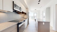 This modern one-bedroom, one-bathroom unit at 280 Montgomery Street in Ottawa offers a thoughtfully designed open-concept layout that maximizes its compact footprint. Light-toned hardwood flooring runs throughout, complementing the bright, white interior and creating a clean, contemporary aesthetic.

The kitchen is a standout feature, equipped with stainless steel appliances including a range, built-in microwave, and dishwasher. Two-tone cabinetry — white upper cabinets paired with taupe lower drawers — is accented by warm wood open shelving and a fine mosaic tile backsplash. A stone-topped island provides additional prep and dining space, while under-cabinet lighting adds a refined touch.

The kitchen flows seamlessly into the combined dining and living area, which leads to a private balcony with views of the Ottawa skyline and surrounding greenery, allowing ample natural light to fill the space.

The bedroom is a calm, well-lit retreat featuring large windows with expansive outdoor views and access to multiple closets, including mirrored sliding door storage.

The bathroom is finished with grey square wall tiles, a floating vanity with stone countertop, an undermount sink, and matte black fixtures. In-unit laundry adds everyday convenience, completing this efficiently designed urban residence.