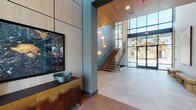 This two-story amenity facility presents a thoughtfully designed modern interior defined by clean architectural lines, warm wood accents, and abundant natural light. The ground floor begins with a dramatic entrance lobby featuring large-format tile flooring, warm wood ceiling panels, and a sculptural wood-and-metal staircase with decorative railings. Adjacent to the entry is a mail and business center with a streamlined wood desk, vibrant velvet seating, and a full bank of individual mailboxes set against a textured accent wall.

A café-style lounge area offers hardwood-style flooring, built-in banquette seating with open shelving, bistro tables, and large windows overlooking the surrounding landscape. A fully equipped demonstration kitchen features a bold teal island with a waterfall countertop, stainless steel appliances, a statement range hood, and modern pendant lighting. A compact service kitchenette with quartz counters and black hardware provides additional functionality.

The second floor features a soaring double-height lounge filled with floor-to-ceiling windows that flood the space with natural light and panoramic outdoor views. A glass-walled corridor with open mezzanine sightlines connects the upper level, enhancing the sense of openness and visual continuity throughout the building.