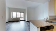 This single-floor space offers a clean, modern aesthetic defined by an open-concept layout that seamlessly connects the kitchen and living areas. The flooring throughout is a rich, dark hardwood that adds warmth and contrast against the crisp white walls and ceilings, creating a polished, contemporary feel.

The kitchen features a prominent peninsula with granite-style countertops, providing ample workspace and a natural division between the cooking and living zones. The cabinetry is finished in white, contributing to the bright and airy atmosphere of the space.

The living area benefits from generous natural light streaming through large, multi-pane windows that frame views of the surrounding urban landscape. A wall-mounted radiator beneath the windows ensures year-round comfort while maintaining a streamlined interior aesthetic.

The open floor plan allows for flexible furniture arrangements, making efficient use of the available space. The clean lines, neutral color palette, and thoughtful layout combine to create an inviting and functional environment. The overall ambiance is modern and understated, well-suited for those who appreciate minimalist design paired with the energy and convenience of an urban setting.