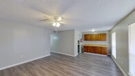 This single-floor unit offers a practical and well-organized layout across several distinct living spaces. The open living and dining area serves as the heart of the unit, featuring durable wood-look laminate flooring, neutral gray-toned walls, crisp white baseboards, a textured popcorn ceiling, and a ceiling fan with integrated lighting. A large window and a front entry door with a sidelight allow natural light to fill the space, creating a bright and airy atmosphere. Adjacent to the living area, a dedicated dining space connects directly to the kitchen, which features a classic galley configuration lined with white cabinetry, gray laminate countertops, a stainless steel sink, and white appliances including a coil-top electric range and a full-size refrigerator. Warm-toned wood cabinetry with yellow laminate countertops is visible in the dining area, providing additional storage and workspace. The unit includes two bedrooms, each finished with the same wood-look flooring, neutral walls, a ceiling fan, and a window for natural light. The full bathroom is clean and functional, outfitted with a white vanity featuring a stone-look countertop, a standard toilet, and a full tub-shower combination with a white surround and chrome fixtures.