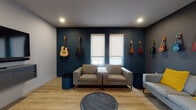 This single-floor music lounge is a thoughtfully designed space that blends creative inspiration with comfortable, modern aesthetics. The room's most striking feature is its deep teal and charcoal accent walls, which serve as a dramatic backdrop for a curated collection of wall-mounted guitars and ukuleles — ranging from acoustic to electric instruments in a variety of colors and styles. The display spans multiple walls, giving the space a gallery-like quality centered around music. Light wood plank flooring runs throughout, providing a warm contrast to the bold wall tones and contributing to the room's inviting atmosphere. Seating is arranged for both comfort and conversation, featuring contemporary grey sofas and armchairs accented with vibrant yellow throw pillows, centered around a round dark wood coffee table. Recessed lighting in the ceiling provides even, ambient illumination throughout. Two windows along the teal accent wall are fitted with roller shades, allowing for adjustable natural light. A wall-mounted flat-screen TV is positioned near the entry door, adding a functional media element to the space. The entry features a glass-panel door with a keypad access system, a wall-mounted sanitizer dispenser, and security hardware, suggesting a semi-public or shared-use environment. Overall, the space offers a vibrant, music-inspired atmosphere ideal for creative gatherings or casual lounging.