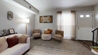 Located on Derrer Road in Columbus, Ohio, this two-story, 2-bedroom, 1-bathroom home offers a warmly curated interior with a cohesive blend of rustic and modern design elements throughout. The main floor hosts a living room, dining room, kitchen, and staircase access to the upper level. The living room is carpeted and inviting, featuring neutral tones, a plush sofa, accent chairs, and decorative wood wall art that adds a rustic character to the space. The dining area is stylishly appointed with a round walnut table, sage green upholstered chairs, a bold floral area rug, and a pair of arched window-pane mirrors that lend architectural interest to the room. The galley-style kitchen is efficiently laid out with dark shaker cabinets, white countertops, black appliances, and wood-look flooring, providing both function and style. Upstairs, the primary bedroom showcases warm hardwood floors, a rattan-style bed frame with coordinating dresser and nightstands, and botanical wall art that creates a serene retreat. The second bedroom is playfully designed with a safari theme, complete with a canopy bed, jungle-print rug, and animal-themed décor. The shared bathroom features a tub-shower combination with a vibrant botanical shower curtain, wood floors, and a rattan wall shelf, giving it a lively, nature-inspired character.