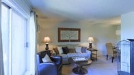 This single-floor space offers a comfortable and thoughtfully arranged living environment. The living room serves as the centerpiece, showcasing a warm, neutral color palette with soft blue-grey walls and a textured ceiling that adds subtle architectural character. Natural light filters in through draped windows, brightening the room and enhancing its inviting atmosphere.

The living area is furnished with a plush, light-toned sofa adorned with an assortment of decorative pillows, complemented by a classic leather armchair in deep navy blue. A round, dark wood pedestal coffee table anchors the seating arrangement, while matching table lamps on either side of the sofa cast a warm ambient glow. A framed botanical wall art piece featuring a tree motif serves as a focal point above the sofa, adding a natural, elegant touch to the room.

The flooring features light-toned carpeting that contributes to the overall warmth and comfort of the space. A small side table beside the sofa provides additional surface space, enhancing the room's functionality. The overall ambiance is calm, classic, and well-balanced, making it a welcoming retreat suited for relaxed everyday living.