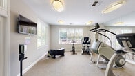 Located at 7030 Gentle Shade Rd in Columbia, MD, this well-appointed fitness center offers a comprehensive workout environment within a single-floor layout. The space is designed with functionality and comfort in mind, featuring neutral gray walls, carpeted flooring, and flush-mount ceiling lights that create a clean, bright atmosphere throughout.

The main exercise area is generously sized, housing a robust selection of cardio equipment including treadmills, a StairMaster, an elliptical trainer, a stationary spin bike, and additional functional fitness accessories such as a training tire. Full-length mirrored walls along one side of the room enhance the sense of space and allow users to monitor their form during workouts. Multiple windows fitted with horizontal blinds invite ample natural light and provide pleasant views of the surrounding greenery.

A wall-mounted flat-screen television is conveniently positioned near the entrance, and a water cooler station offers easy hydration access. The entryway features elegant glass-paneled French doors with transom windows above, opening to a welcoming outdoor area with landscaped surroundings. Sanitization stations are also present near the entrance, reflecting a commitment to cleanliness. Interior glass-paned doors connect areas within the facility, adding an architectural refinement that complements the space's overall polished and accessible design.
