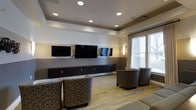 This well-appointed single-floor communal lounge offers a polished, contemporary setting designed for both relaxation and entertainment. The focal point of the room is a sleek, wall-mounted media wall featuring multiple flat-screen televisions arranged side by side, supported by an expansive built-in dark wood cabinet unit that spans the length of the wall, providing ample storage with multiple compartments. The ceiling features a tray design with recessed can lighting that evenly illuminates the space, complemented by warm wall sconces that add a refined ambient glow. The two-tone wall treatment — with a lighter upper half and a deeper grey lower half — adds visual depth and a contemporary aesthetic. Natural light filters in through large windows dressed with neutral drapes on one side of the room, further brightening the interior. The flooring consists of light-toned hardwood-style planks accented by patterned area rugs that tie the space together. Comfortable upholstered club chairs with a circular patterned fabric are thoughtfully arranged throughout, creating an inviting atmosphere suited for gathering, viewing, or casual conversation. Overall, the space exudes a clean, modern character with attention to both function and design.