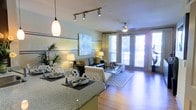 This single-floor space showcases an open-concept layout that seamlessly connects the dining and living areas, creating a cohesive and functional flow throughout. The dining area features a granite countertop bar with elegant place settings and a stylish pendant light overhead, making it an ideal spot for casual meals or entertaining. Track lighting along the ceiling provides targeted illumination, complementing the warm glow of table lamps placed throughout the space.

The living area is anchored by hardwood flooring and a patterned area rug, adding texture and warmth underfoot. A ceiling fan is centrally positioned, enhancing comfort while contributing to the room's modern aesthetic. The walls are painted in soft, neutral tones and adorned with artwork that adds a sophisticated touch. Lush indoor plants introduce a natural, refreshing element to the interior.

Large floor-to-ceiling windows with sheer drapes line the far wall, allowing abundant natural light to pour in and offering a connection to the outdoors. The overall ambiance is bright, contemporary, and welcoming, with a cohesive design that balances style and livability throughout.