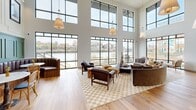 Located at 451 W Blount Ave in Knoxville, Tennessee, this generously scaled two-story amenity space delivers an impressive blend of industrial character and contemporary design. The main floor commands attention with dramatic double-height ceilings lined by floor-to-ceiling black-framed windows that frame sweeping views of the waterfront and Knoxville skyline. Warm natural light floods the space across wide-plank hardwood flooring throughout. A striking exposed brick ceiling anchors one section of the main level, lending historic texture that contrasts beautifully with the crisp white walls. This area features a cozy lounge with a central fireplace, a pool table, and casual seating. A fully equipped kitchen showcases dark granite countertops, rich wood cabinetry, golden zellige tile backsplash, open shelving, and stainless steel appliances. A sophisticated dining lounge area features built-in leather banquette seating beneath paneled sage-green wainscoting and pendant lighting. Two staircases lead to the upper level, a bright, airy lounge bathed in natural light from large windows overlooking the surrounding neighborhood. This secondary level offers inviting seating arrangements beneath a stylish shiplap ceiling with designer light fixtures, completing an overall atmosphere that is warm, curated, and community-focused.