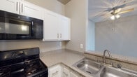 This single-floor residence in Santa Clarita, California, offers a thoughtfully arranged layout with distinct living zones visible in the overhead dollhouse view. The kitchen is a functional and well-appointed space, featuring white shaker-style cabinetry with brushed nickel hardware, speckled granite-look countertops, a stainless steel double-basin sink, and black appliances including a gas range and over-the-range microwave. A ceiling fan with light fixture is positioned in the adjoining area, providing both airflow and illumination. The kitchen opens toward neighboring living spaces, creating a connected feel throughout.

The living and bedroom areas are finished with plush neutral carpeting and painted in warm greige tones, contributing to a calm, cohesive atmosphere. Tall ceilings in the main living area lend a sense of openness, with vertical blinds covering a large sliding glass door that introduces natural light. Additional rooms feature corner window configurations, further brightening the interiors with natural daylight and views of surrounding greenery.

The bathroom is finished with rich wood-look flooring, white cabinetry, a granite-style countertop, a large mirror, and a shower-tub combination, offering a clean and practical design. Overall, the space presents a comfortable and well-maintained environment.