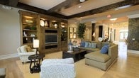 This single-floor space exudes a refined, welcoming ambiance anchored by a thoughtfully designed lounge or common area. The centerpiece of the room is a floor-to-ceiling built-in entertainment unit crafted from rich, dark wood, featuring open shelving for decorative accents and a recessed space for a flat-screen television. The ceiling draws the eye upward with its elegant coffered design, accented by bold dark wood beams that add architectural depth and contrast against the warm neutral tones of the walls and flooring. Decorative columns further define the space, lending a classical architectural touch. The seating arrangement is both generous and inviting, with a large sectional sofa dressed in earthy tones and accented with blue and teal throw pillows, complemented by additional accent chairs upholstered in a soft patterned fabric. A substantial dark wood coffee table anchors the seating area, while tasteful table lamps and a large indoor plant introduce warmth and life to the room. Recessed lighting throughout ensures the space is well-lit, enhancing its sophisticated yet comfortable character. Overall, the space strikes a balance between elegance and approachability, making it ideal for relaxation and social gatherings.