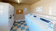 This shared laundry facility is a practical, single-floor space designed to serve the everyday needs of its users. The room is immediately recognizable by its distinctive blue and yellow checkered vinyl tile flooring, which adds a touch of character to the otherwise utilitarian environment.

The facility is outfitted with a row of commercial-grade top-load washing machines by Speed Queen, lined neatly along one wall and managed through a CSC payment service system. Adjacent to the washers, a set of stacked dryer units maximizes the use of vertical space, providing ample drying capacity. CSC service information is posted on the walls in both English and Spanish for easy reference.

A generously sized folding counter with a blue laminate surface runs along one wall beneath a large window, which allows natural light to fill the space and offers a view of the surrounding parking area and neighboring buildings. This counter provides a convenient surface for sorting and folding laundry.

A cork bulletin board near the entrance serves as a communication hub for notices and announcements. Overhead fluorescent lighting ensures the room is well-lit throughout. The space is accessed through a solid six-panel door equipped with a door closer. A baseboard heater along the wall provides warmth during colder months, making this facility a comfortable and fully functional resource.