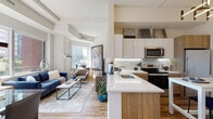 Located in the heart of White Plains, NY, this modern residence at 1 Martine Ave offers a thoughtfully designed open-concept floor plan that seamlessly connects the living, dining, and kitchen areas. The kitchen is outfitted with white upper cabinetry, warm wood-toned lower cabinets, a textured tile backsplash, stainless steel appliances, and a sleek white island that doubles as a casual dining space. The adjacent living and dining area is bathed in natural light through large floor-to-ceiling windows, revealing sweeping views of the White Plains skyline and surrounding greenery. Exposed ceiling beams add architectural character to the bright, airy living space. Hardwood flooring runs throughout the main living areas, contributing to a warm and cohesive aesthetic. The primary bedroom features wide windows with city views, hardwood floors, and generous wall space, while the second bedroom offers a similarly comfortable and well-lit retreat. Two full bathrooms are finished with large-format white tile, wood-toned vanities, and clean white countertops, conveying a crisp, contemporary feel. The overall design balances modern finishes with a welcoming ambiance, making this a refined urban living option in downtown White Plains.