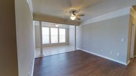 This single-story space showcases a thoughtfully designed open layout that seamlessly connects two distinct living areas through a pair of decorative white columns. The columns serve as an elegant architectural divider, delineating the transition between a section finished with rich, dark hardwood flooring and a lighter tile-floored area near the front of the space. The neutral gray walls are complemented by crisp white crown molding and wide baseboards, contributing to a clean and polished aesthetic throughout.

Each zone is equipped with its own ceiling fan with integrated lighting, ensuring both comfort and practicality across the full expanse of the space. A large multi-pane window at the front of the tile area allows generous natural light to fill the room, brightening the interior and creating an airy, welcoming atmosphere. The textured ceiling adds subtle visual depth overhead.

The overall ambiance is calm and neutral, offering a versatile canvas well-suited to a variety of furnishing styles. The combination of contrasting floor materials and architectural column details gives the space a sense of character and intentional design.