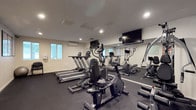 This fitness center is a fully outfitted single-floor facility designed to support a well-rounded workout routine. The main exercise area is furnished with multiple cardio machines, including treadmills, ellipticals, and stationary bikes, all positioned to maximize use of the available floor space. A dedicated strength training machine provides options for resistance-based workouts, while a stability ball and seating area offer a spot for cooldowns and flexibility work.

The space is finished with dark rubber flooring throughout, providing both durability and safety underfoot. Recessed lighting illuminates the room evenly, complemented by natural light filtering in through several frosted windows. A wall-mounted mini-split HVAC unit ensures a comfortable climate year-round.

Large wall mirrors on one side of the room expand the sense of space and allow users to monitor their form during exercise. Multiple wall-mounted flat-screen TVs are strategically placed for viewing while using the cardio equipment. Educational fitness and exercise stretch posters are displayed along the walls, offering useful guidance for users of all experience levels.

A separate adjacent room, accessible through a frosted glass door, provides additional space, along with a hand sanitizer dispenser and workout schedule boards, reinforcing the facility's clean and organized environment.