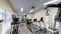 This fitness center offers a comprehensive workout environment designed to meet a variety of exercise needs. The space is outfitted with multiple cardio machines, including treadmills and an elliptical trainer from TRUE Fitness, providing residents with quality equipment for aerobic training. A dedicated free weights area features a full dumbbell rack, while a multi-station strength training machine supports a wide range of resistance exercises. Full-length mirrors line the walls, creating a sense of openness and allowing users to monitor their form during workouts. The polished concrete flooring is both durable and easy to maintain, well-suited for a high-traffic fitness environment. Bright flush-mounted ceiling lights ensure the space is thoroughly illuminated throughout the day. Ventilation is supported by ceiling air vents, keeping the environment comfortable during intense sessions. A mounted television is also present, offering entertainment while exercising. The neutral color palette of white and gray walls contributes to a clean, focused atmosphere. Overall, this fitness center is thoughtfully arranged to maximize functionality, providing a well-rounded facility for cardiovascular, strength, and general fitness training.