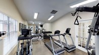 This fitness center offers a comprehensive workout environment designed to meet a variety of exercise needs. The space is outfitted with multiple cardio machines, including treadmills and an elliptical trainer from TRUE Fitness, providing residents with quality equipment for aerobic training. A dedicated free weights area features a full dumbbell rack, while a multi-station strength training machine supports a wide range of resistance exercises. Full-length mirrors line the walls, creating a sense of openness and allowing users to monitor their form during workouts. The polished concrete flooring is both durable and easy to maintain, well-suited for a high-traffic fitness environment. Bright flush-mounted ceiling lights ensure the space is thoroughly illuminated throughout the day. Ventilation is supported by ceiling air vents, keeping the environment comfortable during intense sessions. A mounted television is also present, offering entertainment while exercising. The neutral color palette of white and gray walls contributes to a clean, focused atmosphere. Overall, this fitness center is thoughtfully arranged to maximize functionality, providing a well-rounded facility for cardiovascular, strength, and general fitness training.