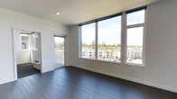 This contemporary single-floor unit showcases a clean, modern design characterized by crisp white walls, recessed lighting, and rich dark hardwood flooring that runs throughout the space. The open main living area is a standout, bathed in an abundance of natural light courtesy of expansive floor-to-ceiling windows that frame sweeping views of neighboring residential buildings and a lush backdrop of evergreen trees. A sliding glass door leads directly to a private balcony, extending the living space outdoors and enhancing the connection to the surrounding scenery. The layout flows naturally into a secondary room, visible through an open doorway, which benefits from its own window and receives generous natural light. A partial view of cabinetry suggests a kitchen or utility area along one edge of the main room. The overall ambiance is bright, airy, and minimalist, with a neutral color palette that complements the dark flooring and creates a polished, cohesive aesthetic. The elevated position of the unit further enhances the panoramic outdoor views, making natural light and scenery central features of this well-appointed space.