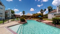 This amenity space combines an inviting outdoor pool area with a well-appointed fitness center, creating a comprehensive recreational destination. The outdoor pool features clear, turquoise water set within a spacious surround finished in brick-accented hardscaping. Raised planter beds with tropical plantings add a lush aesthetic, while a covered gazebo with seating provides a shaded retreat. Lounge chairs and a patio umbrella complete the outdoor setting, all framed by mature trees and manicured landscaping under open skies.

The fitness center is housed in a bright, airy interior characterized by soaring ceilings and abundant natural light streaming through multiple large windows. Patterned carpet tiles in blue and grey tones cover the floor throughout. The space is thoughtfully divided into functional zones: one area features a power rack, cable machine, rowing machine, and weight bench for strength training, while another section is dedicated to functional fitness equipment, including a multi-station cable rig, medicine balls in assorted weights, battle ropes, stability balls, and a full dumbbell rack with floor-to-ceiling mirrors. A dedicated cardio alcove houses treadmills and a Precor cycle, accompanied by a wall-mounted TV. Built-in shelving near the entrance offers additional storage and supplies.