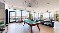Located at 25 Maple Ave in New Rochelle, NY, this contemporary amenity lounge offers a generously sized, single-floor layout designed for both relaxation and recreation. Floor-to-ceiling windows wrap around multiple zones of the space, flooding the interior with natural light and framing panoramic views of the New Rochelle skyline and surrounding cityscape. A dedicated outdoor terrace, accessible via large glass doors, extends the living area and provides an open-air setting furnished for dining and socializing.

Inside, the lounge is organized into distinct yet interconnected areas. A central games zone showcases a full-sized pool table anchored beneath a striking sculptural pendant light, with curved green leather banquette seating adding a bold retro-modern accent. Adjacent to this is a comfortable lounge area furnished with contemporary sofas, patterned area rugs, and curated coffee tables. A media corner features a large wall-mounted television set against calming blue-toned walls. The kitchen bar area offers counter seating with warmly finished stools. Throughout, light wood herringbone flooring, sage green accent walls, and thoughtfully placed greenery create a cohesive, inviting ambiance suitable for community gatherings or quiet downtime.