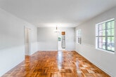 This single-floor residence is defined by its striking parquet hardwood flooring, which flows consistently throughout the living, dining, and bedroom areas, lending warmth and visual character to each room. The open living and dining areas are connected seamlessly, with chair rail molding running along the walls adding a classic architectural touch. A chandelier anchors the dining area, while large windows allow natural light to filter in, creating a bright and welcoming atmosphere.

The spacious living room features multiple windows, a wall-mounted air conditioning unit, and a radiator, ensuring year-round comfort. A ceiling fan with integrated lighting is present in each bedroom, complementing the natural light provided by outward-facing windows. One bedroom includes a built-in closet with shelving, offering practical storage. Radiators are present throughout, consistent with the building's traditional heating system.

The kitchen features warm-toned wood cabinetry, dark countertops, stainless steel appliances including a full-size refrigerator, and a classic black-and-white diamond-patterned tile floor. Two windows bring in natural light. The bathroom is finished with white tile, a radiator, a window with outdoor views, and a glass-door shower enclosure, maintaining the home's clean, classic aesthetic throughout.