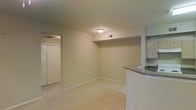 This single-floor residential space offers a clean, functional layout designed for comfortable everyday living. The main living area is finished with wall-to-wall carpeting in a neutral tone, contributing to a warm and quiet atmosphere throughout. Smooth, light-colored walls and standard-height ceilings give the space a bright, open feel, with a flush-mount ceiling light providing even illumination.

A curved kitchen counter with a dark laminate surface extends into the living area, creating a natural pass-through that connects the kitchen and living spaces. Above the counter, open shelving built into the wall offers convenient storage and display space. This architectural detail adds a subtle design element while enhancing the practicality of the layout.

A doorway leads to an adjacent room or hallway, alongside a closet fitted with classic bifold doors — providing ample storage within the living area itself. Ceiling vents and a wall-mounted thermostat indicate a central HVAC system for year-round climate control.

The overall palette — beige carpet, white walls, and white trim — creates a neutral, move-in-ready environment. The straightforward, efficient design of the space makes it well-suited for a variety of living arrangements, with thoughtful built-in features that maximize usability.