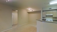 This single-floor residential space offers a clean, functional layout designed for comfortable everyday living. The main living area is finished with wall-to-wall carpeting in a neutral tone, contributing to a warm and quiet atmosphere throughout. Smooth, light-colored walls and standard-height ceilings give the space a bright, open feel, with a flush-mount ceiling light providing even illumination.

A curved kitchen counter with a dark laminate surface extends into the living area, creating a natural pass-through that connects the kitchen and living spaces. Above the counter, open shelving built into the wall offers convenient storage and display space. This architectural detail adds a subtle design element while enhancing the practicality of the layout.

A doorway leads to an adjacent room or hallway, alongside a closet fitted with classic bifold doors — providing ample storage within the living area itself. Ceiling vents and a wall-mounted thermostat indicate a central HVAC system for year-round climate control.

The overall palette — beige carpet, white walls, and white trim — creates a neutral, move-in-ready environment. The straightforward, efficient design of the space makes it well-suited for a variety of living arrangements, with thoughtful built-in features that maximize usability.
